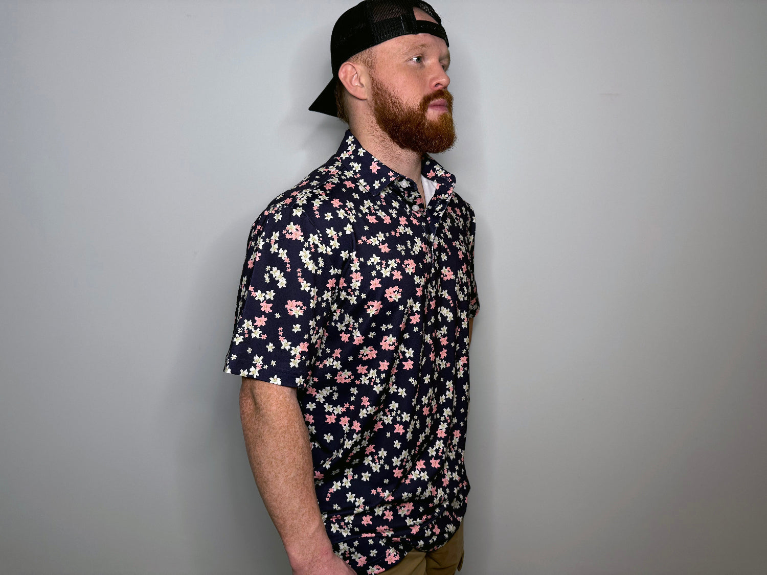 A man wearing a navy polo shirt with a floral design, featuring pink and white petals on a dark background, and a black cap.