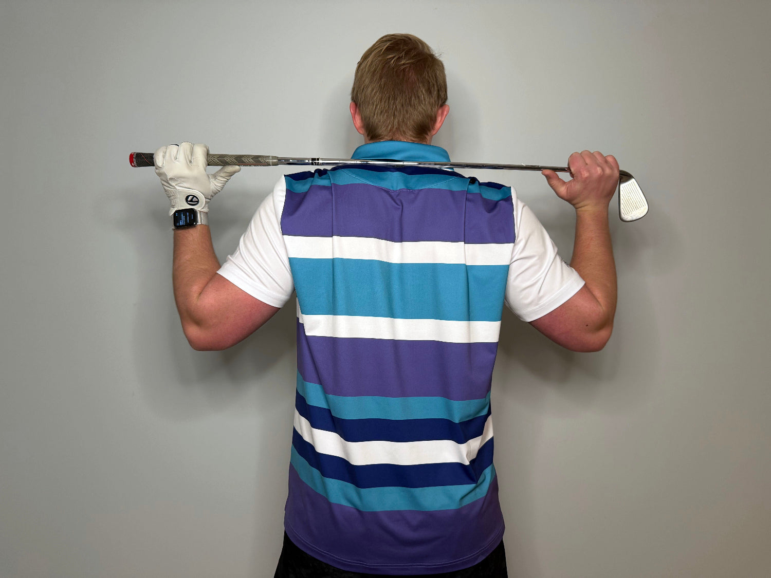 Man standing with a golf club over his shoulders wearing a purple white blue and teal striped polo