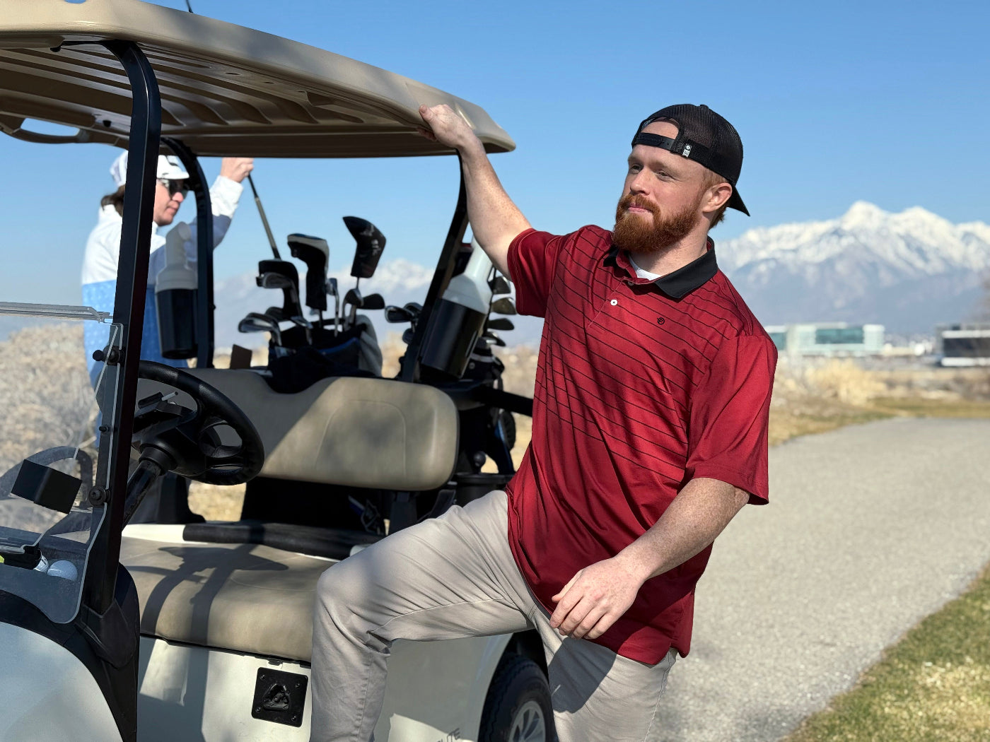 A man wearing a red polo shirt with a white collar, sitting on a golf cart with a mountainous landscape in the background.