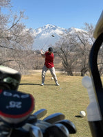 A person playing golf, wearing a red polo shirt, with the golf club in mid-swing. The background includes a mountain range, and a clear sky.