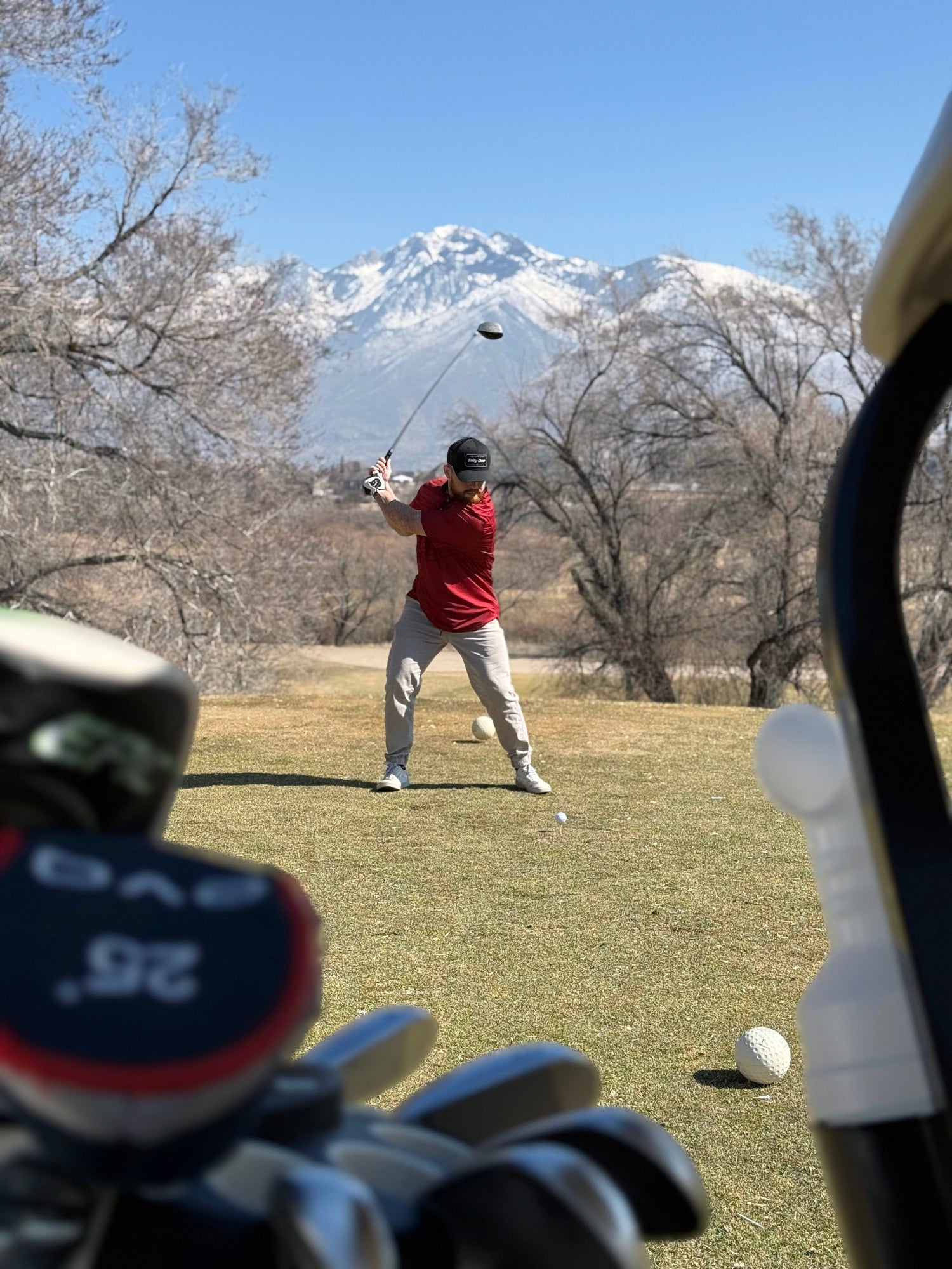 A person playing golf, wearing a red polo shirt, with the golf club in mid-swing. The background includes a mountain range, and a clear sky.