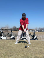 A man in red polo setting up to hit a tee shot with golf cars in the back ground. 
