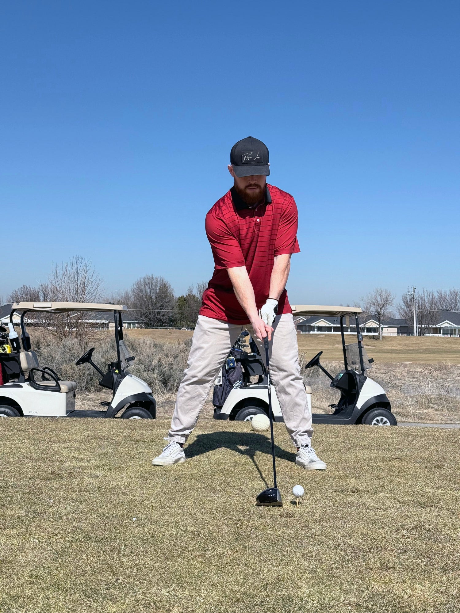 A man in red polo setting up to hit a tee shot with golf cars in the back ground. 