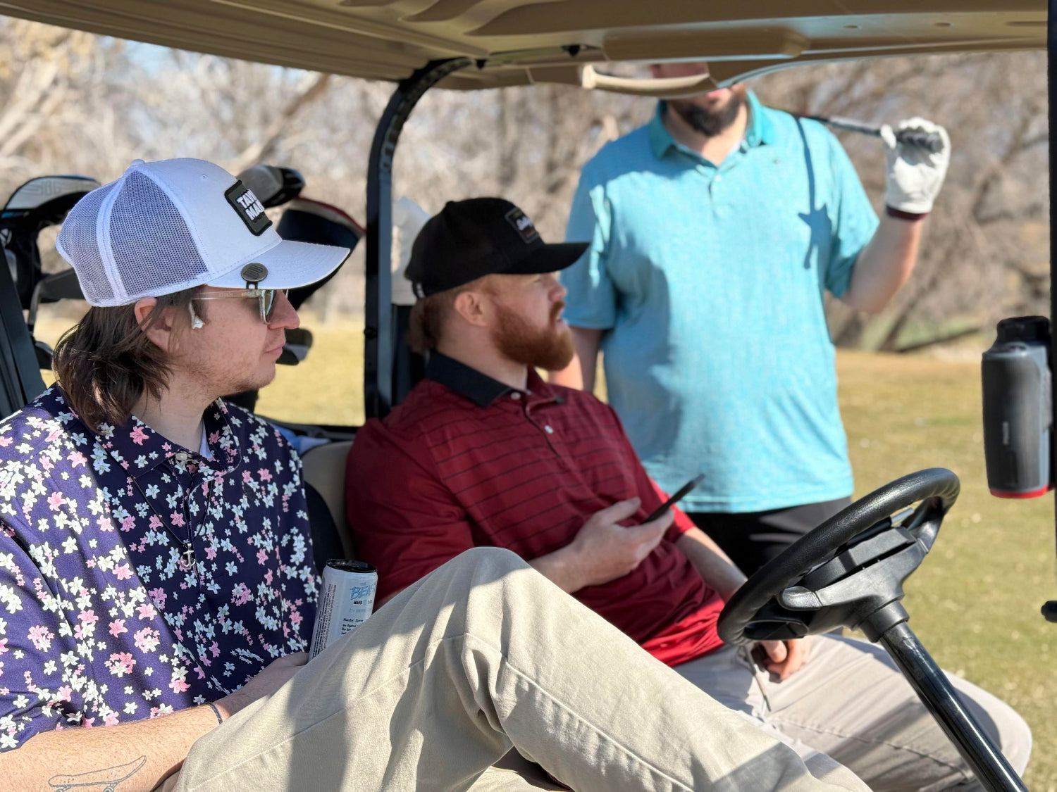 Two men sit in a golf cart with a third man in the back ground