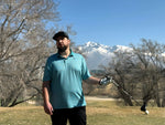 A man standing with a golf driver in his hand. Mountains and trees in the background