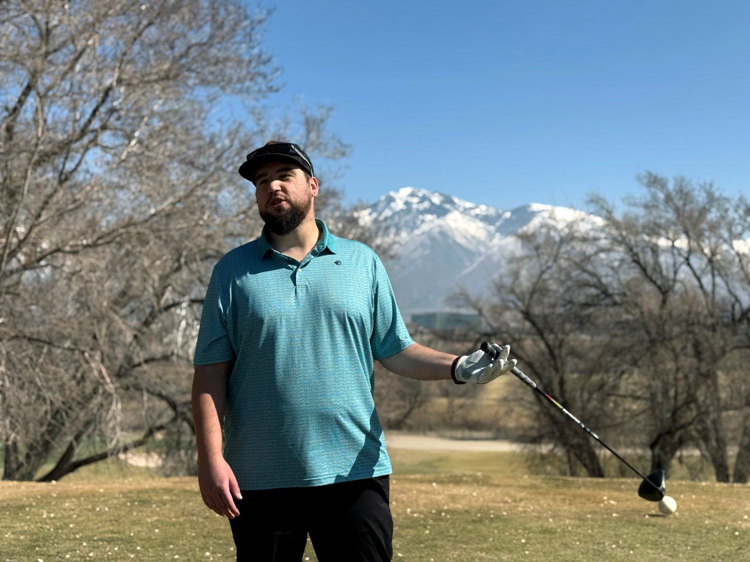 A man standing with a golf driver in his hand. Mountains and trees in the background