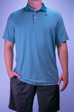 Person wearing a blue patterned polo shirt and black shorts against a purple background