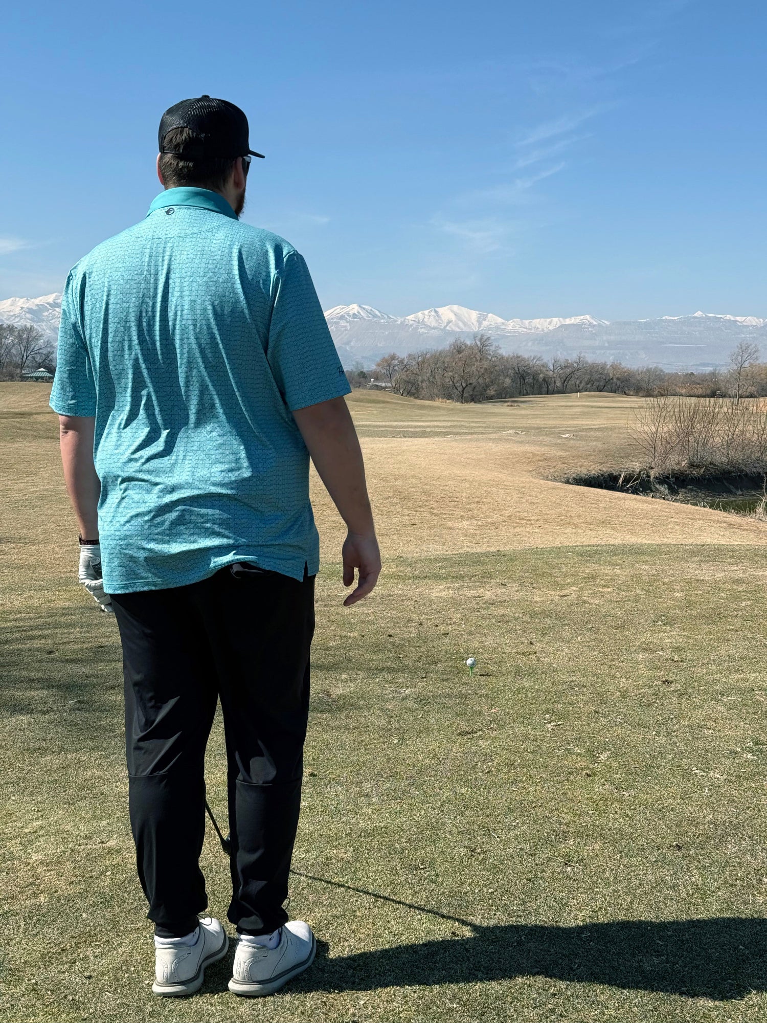 Golfer standing close with blue shirt and black pants. Mountains afar