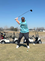 Golfer wearing a blue polo swinging a driver to hit a golf ball. Golf carts in the background