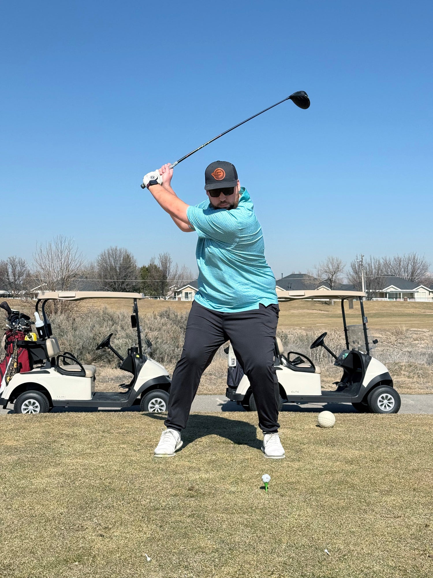 Golfer wearing a blue polo swinging a driver to hit a golf ball. Golf carts in the background