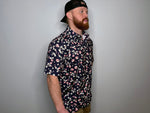 A man wearing a navy polo shirt with a floral design, featuring pink and white petals on a dark background, and a black cap.