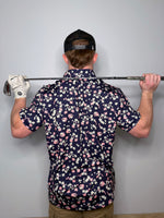 Man wearing a polo with white and pink flowers with dark base. Holding a golf club across his shoulders