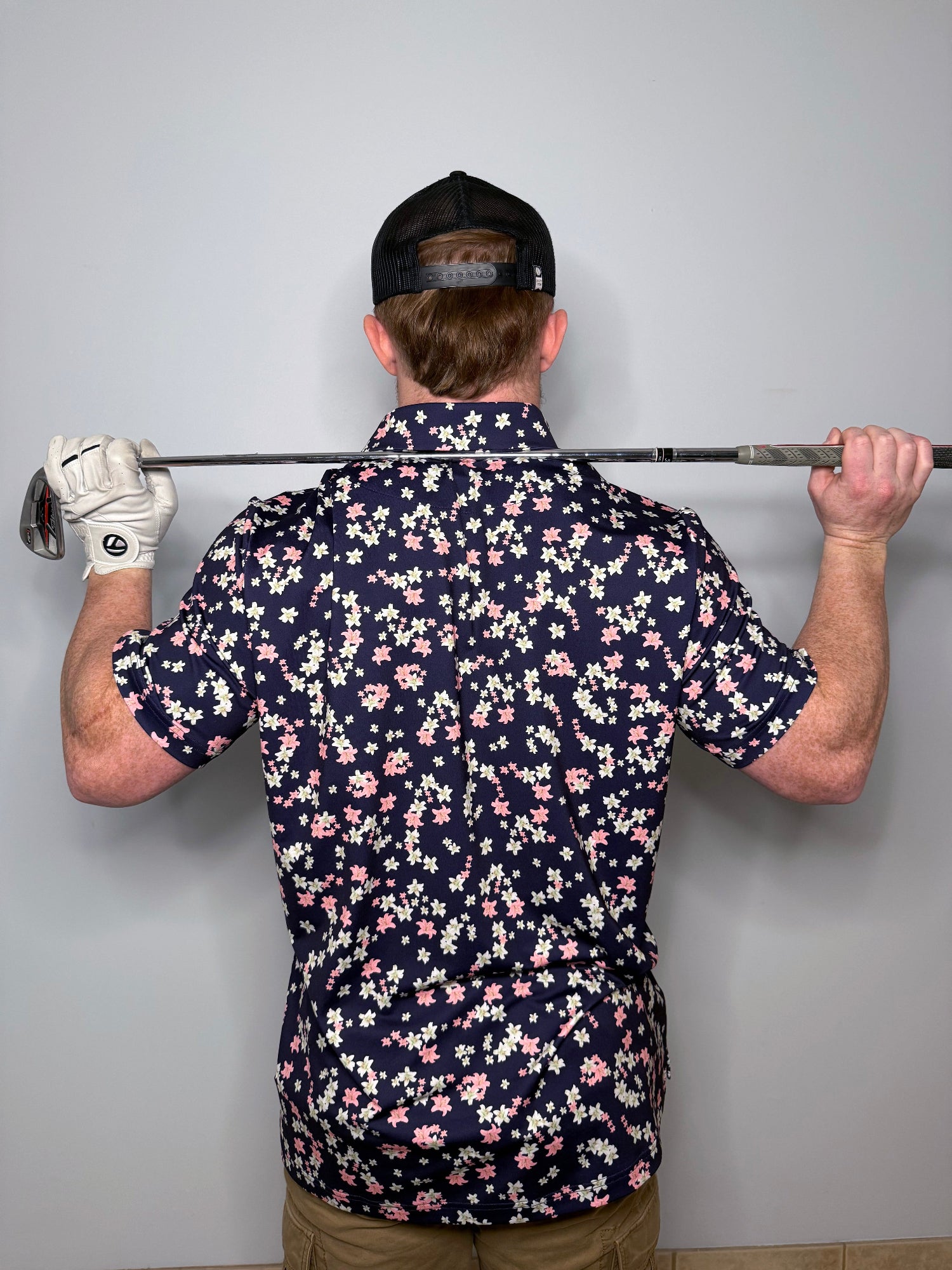 Man wearing a polo with white and pink flowers with dark base. Holding a golf club across his shoulders
