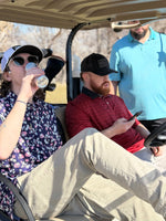 1 man drinking from a can while sitting in a golf cart with another man. Third man standing behind them