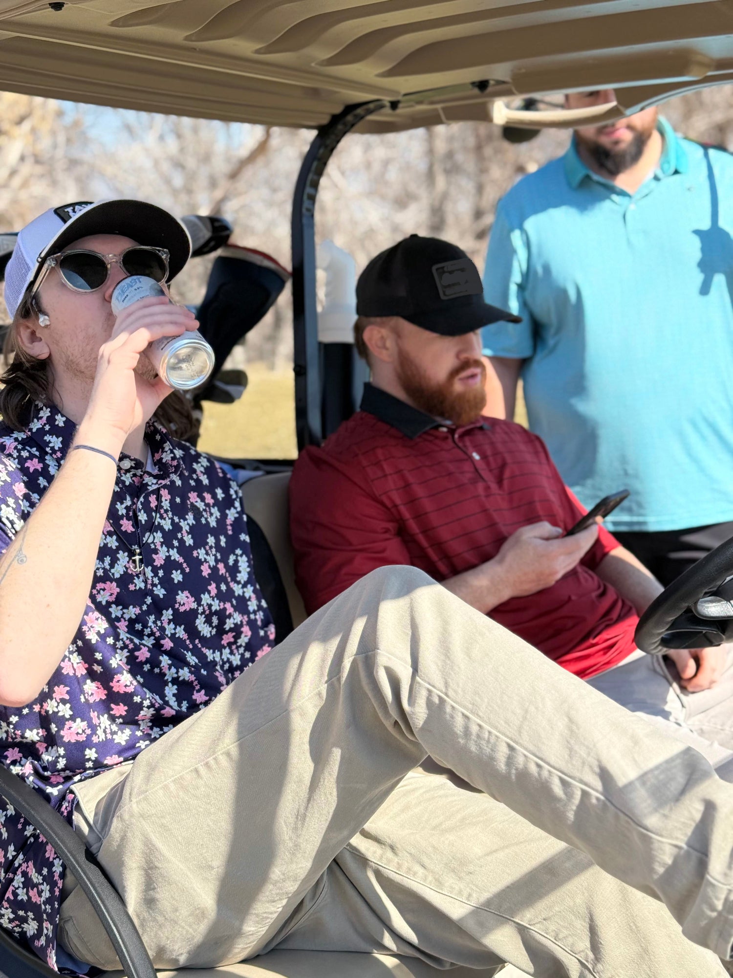 1 man drinking from a can while sitting in a golf cart with another man. Third man standing behind them