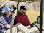 Two guys sitting in a golf cart looking away from the camera. Golf Clubs in the back ground